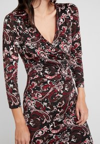 Kvinna i en långärmad svart klänning med ett röd- och vitmönstrat paisleymönster, V-ringning och draperad tyg vid midjan.