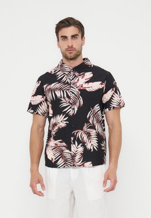 Muž nosi čiernu košeľu s krátkym rukávom s veľkým béžovým a červeným tropickým listovým vzorom, v kombinácii s bielymi nohavicami, stojí na bielom pozadí.