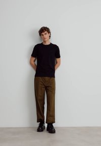 Sort kortærmet t-shirt, olivengrønne bukser og sorte snørebåndsstøvler. Ung mand står foran en neutral baggrund. Casual pasform.