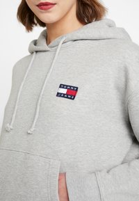 Sudadera con capucha gris con bolsillo frontal, cordones y parche de logo bordado en azul marino con detalles en rojo y blanco. Tela suave y texturizada.