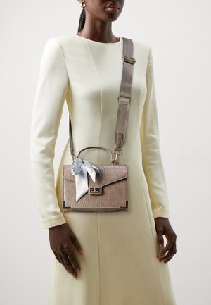 Mujer con un vestido largo de manga larga color crema que lleva un bolso taupe con relieve de cocodrilo, una correa ancha y un detalle de lazo plateado en el frente.