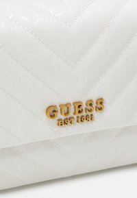 Vit quiltad handväska med präglat monogrammönster, guldfärgade detaljer och "GUESS EST 1981"-logotyp i fet stil.
