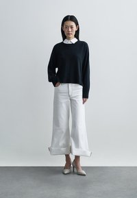 Maglione nero a maglia con orlo a coste, camicia bianca con colletto sotto, abbinati a jeans bianchi a gamba larga e scarpe con tacco argento. Design semplice e pulito.