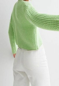 Personne portant un pull en maille texturée vert clair et un pantalon blanc taille haute, vue de dos et de côté, sur un fond uni.