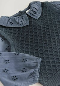 Pletený sveter v tmavozelenej farbe s textúrovaným diamantom, s svetlomodrým vyšívaným golierom a nafúknutými rukávmi.