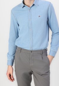 Světle modrá denimová košile s límečkem na knoflíky, dlouhými rukávy a jemným značkovým logem. Šedé kalhoty s hladkým předním dílem a bočními kapsami.