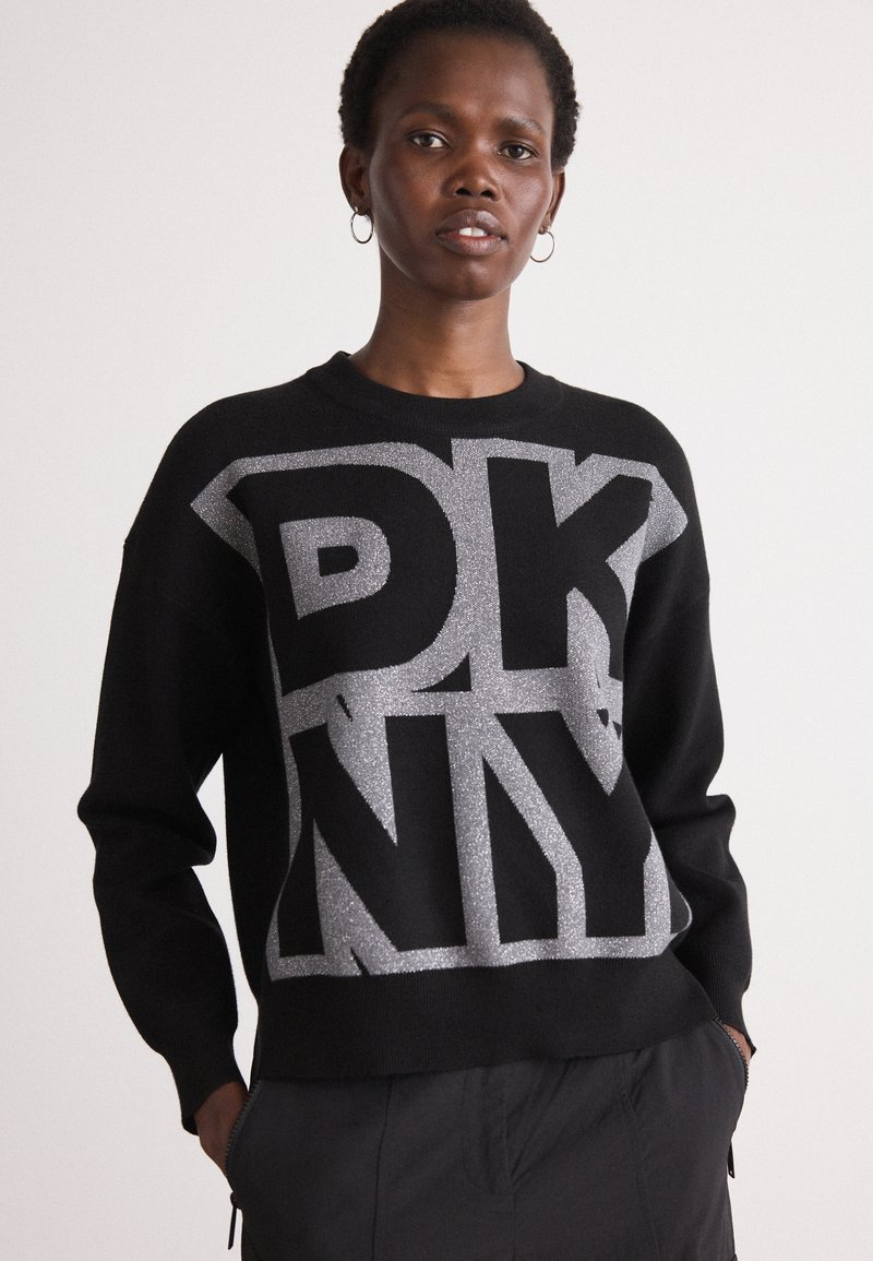 Melnā džemperī ar lielu sudraba "DKNY" grafiku, apaļu kakla izgriezumu un garām piedurknēm. Materiāls šķiet mīksts un ar patīkamu tekstūru.