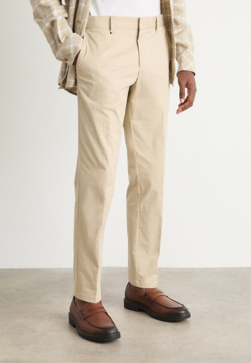 Homme portant un pantalon beige sur mesure, des mocassins en cuir marron, une chemise blanche et un blazer beige à carreaux, debout sur un sol gris.