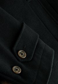 Schwarzer Wollmantel aus strukturiertem Stoff, mit Reverskragen und zwei großen braunen Knöpfen mit vier Löchern. Maßgeschneiderte Gestaltung mit klaren Linien.