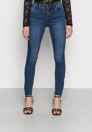 Vrouw die donkerblauwe skinny jeans draagt, zwarte sandaaltjes met hoge hakken en bandjes, en een zwart shirt met lange mouwen en een wit gestippeld patroon.