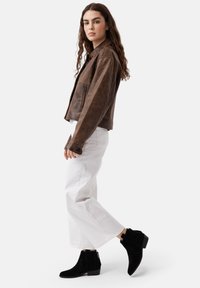 Jachetă din piele maro cu un design ușor scurtat, asortată cu pantaloni albi evazați și cizme negre din suede până la gleznă, cu fermoare laterale.