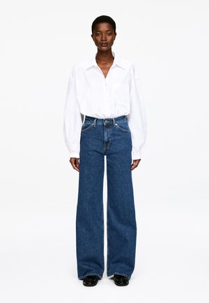 Frau steht und trägt ein lockeres weißes Hemd mit Knöpfen, das in hochtaillierte, weitbeinige blaue Jeans gesteckt ist, sowie schwarze Schuhe.