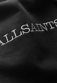 Černá látka s vyšitým nápisem "ALLSAINTS" v bílé barvě. Textura je hladká s mírným pružením, což zvýrazňuje čistý design.