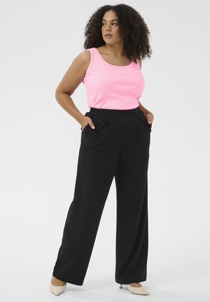 Kvinde med krøllet hår iført en lyserød ærmeløs top, sorte bukser med vide ben og beige spidse hæle, stående med hænderne i lommerne.