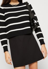 Un pull rayé noir et blanc avec une texture côtelée, associé à une jupe noire unie. Un sac à main en vernis noir avec une forme incurvée et une bandoulière.
