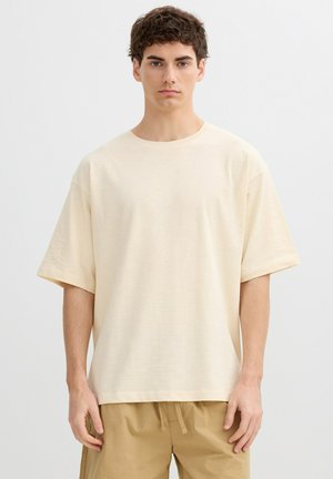 Joven de pie, vistiendo una camiseta beige holgada y pantalones de cuerda color canela, contra un fondo gris claro liso.