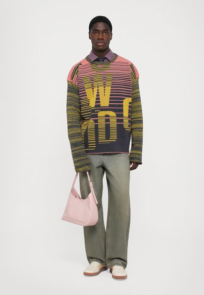 Young man stands wearing oversized striped sweater with large yellow letters, loose gray jeans, white sneakers, holding pale pink handbag.