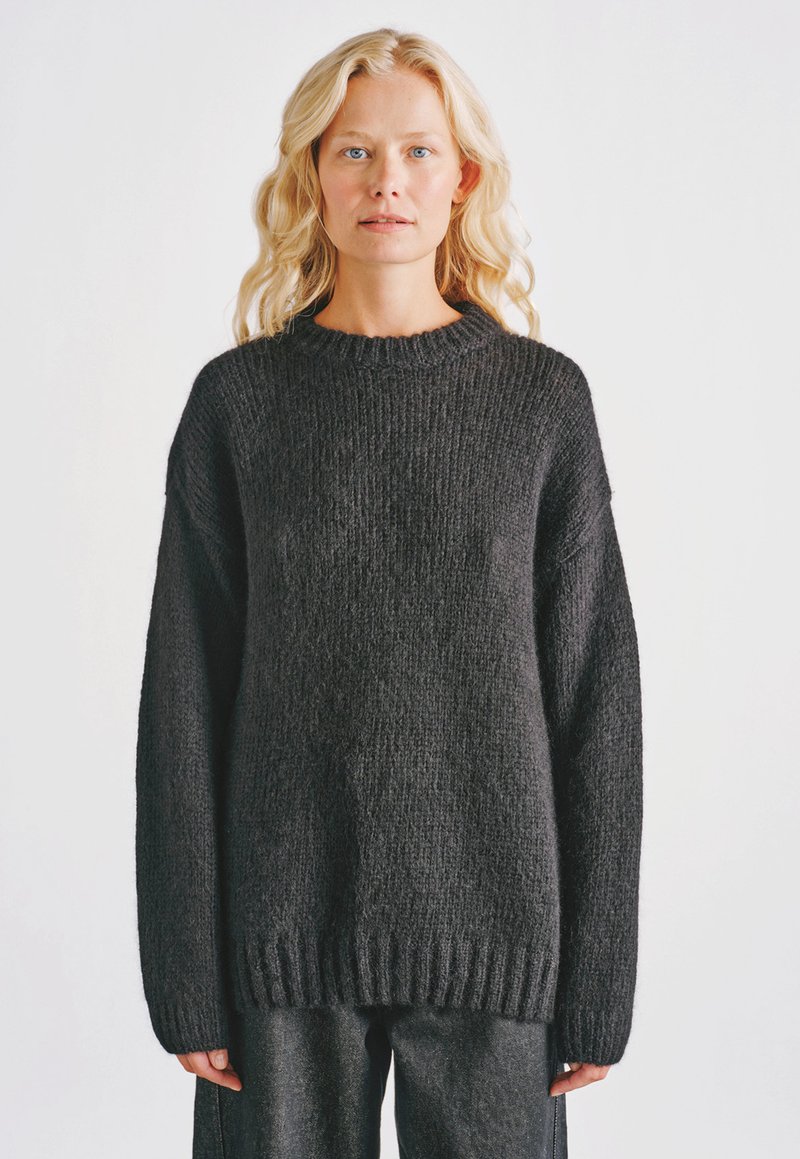 Blondynka o falowanych włosach ubrana w oversize'owy ciemnoszary sweter z dzianiny i dopasowane spodnie, stoi na tle jednolitego jasnego tła.