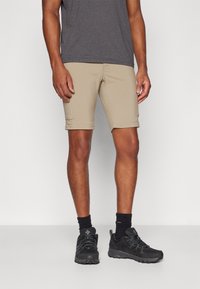 Bežid lühikesed püksid, mis on slim fit stiilis, valmistatud kergest materjalist, külgedel on taskud ja alläär ulatub keskmise reieni, koos mustade tennisetega.