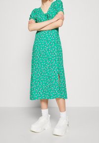 Zielona sukienka w kwiaty z krótkimi bufkami, dekoltem w serek, bocznym rozcięciem i długością do połowy łydki. Połączona z białymi masywnymi sneakersami.