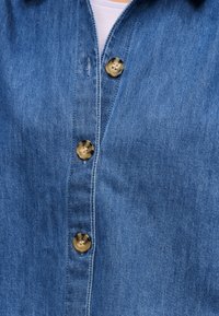 Τζιν πουκάμισο με τρία κουμπιά από χελώνα κλεισμένα πάνω από ένα ανοιχτό μωβ μπλουζάκι.