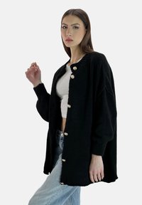 Cardigan negro de corte holgado, tejido texturizado, escote redondo y botones dorados. Combinado con una camiseta blanca y jeans azul claro.