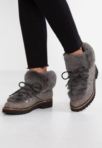 Bottines grises à la cheville avec bord en fausse fourrure, dessus texturé avec des accents cloutés, bout rond et semelle plate. Les lacets noirs complètent le design.