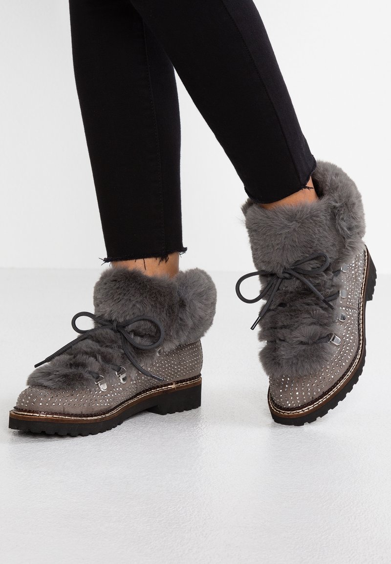 Bottines grises à la cheville avec bord en fausse fourrure, dessus texturé avec des accents cloutés, bout rond et semelle plate. Les lacets noirs complètent le design.