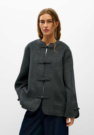 Femme portant une veste en laine gris foncé avec fermetures à boutons à bascule et manches larges, associée à un pantalon bleu foncé, sur fond blanc.
