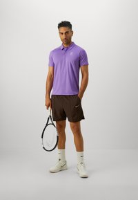Man står i en lila pikétröja, bruna shorts, vita sneakers och strumpor, och håller en tennisracket mot en plain bakgrund.