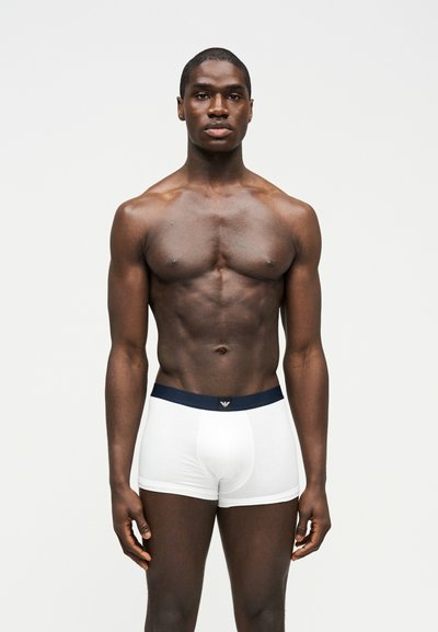 Μυώδης άντρας που στέκεται μπροστά σε λευκό φόντο, φοράει λευκά ανδρικά εσώρουχα με σκούρα μέση και λογότυπο στο κέντρο μπροστά.