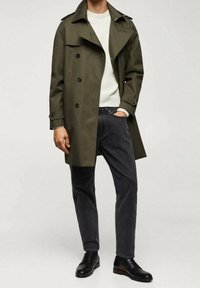 Olivengrüner Trenchcoat mit doppelt-knöpfigem Design, schwarzen Knöpfen und Schulterklappen, getragen über einem weißen Kabelstrickpullover und schwarzen Jeans.