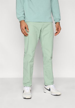 Svetlozelené chinos nohavice s priliehavým strihom a hladkým povrchom, kombinované s bielymi športovými teniskami. Jednoduchý dizajn s minimálnymi detailmi.