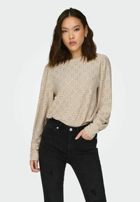 Beige langærmet top med hulmønster, rund halsudskæring og afslappet pasform, parret med højtaljede sorte jeans. Blød tekstur, afslappet stil.