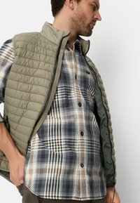 Lichtgewicht doorgestikte bodywarmer in olijfgroen, gecombineerd met een geruit overhemd met knopen in blauwtinten, beige en oranje accenten.