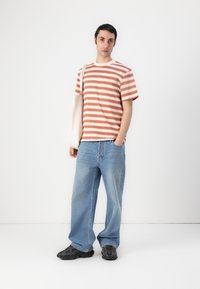 Striped t-shirt in peach and white, relaxed fit; paired with light blue baggy jeans and black sneakers. Model holds a beige tote bag.