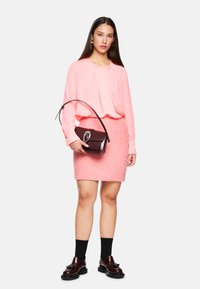 Rosa Bluse mit drapiertem Vorderteil, strukturierter rosa Minirock und dunkelroter Handtasche. Geschlossene Oxford-Schuhe mit dicker schwarzer Sohle und Socken bis zum Knöchel.