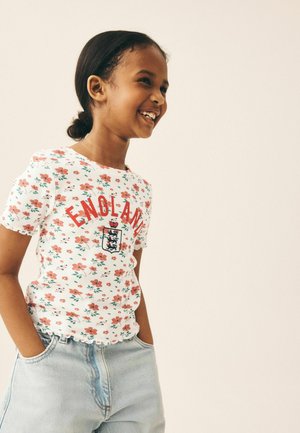 Smilende pige med mørkt hår i en knold iført en hvid blomster-England-shirt og lyseblå jeans med hænderne i lommerne, vender mod højre.