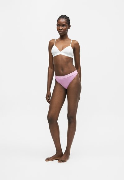 Γυναίκα όρθια ξυπόλυτη, φοράει λευκό bralette και ροζ εσώρουχα με λωρίδα μέσης με λογότυπο πάνω σε απλό λευκό φόντο.