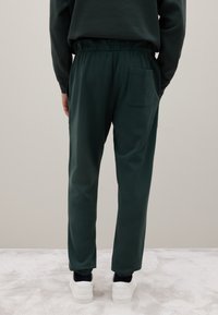 Donkergroene sweatpants van zacht materiaal, met een elastische tailleband, taps toelopende pijpen en een enkele achterzak.