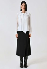 Camicia bianca con dettaglio plissettato e accenti dorati, abbinata a una gonna nera plissettata e stivali neri al ginocchio. Tessuto liscio e design minimalista.