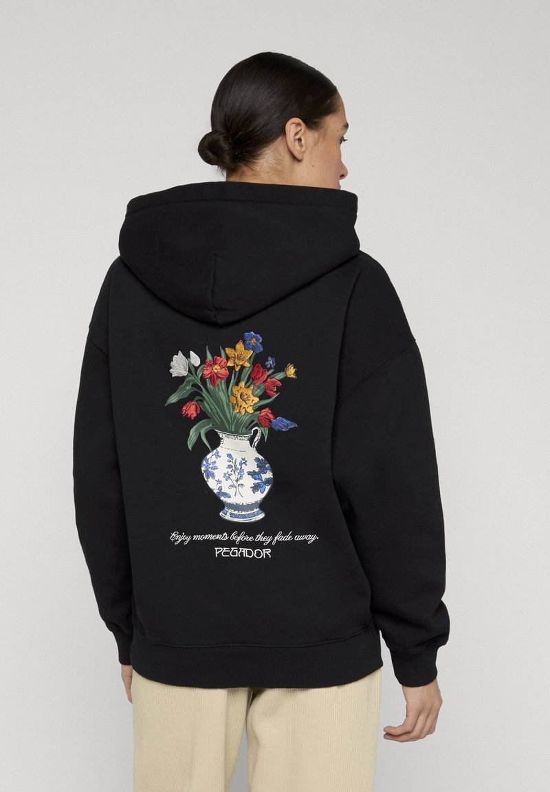Mujer con sudadera negra con capucha con diseño de jarrón floral y texto "Disfruta los momentos antes de que se desvanezcan" en la espalda, de pie frente a un fondo liso.