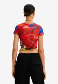 Sopiva lyhythihainen paita, joka esittelee punaisen ja sinisen abstraktin kuvion, kerättyjä sivuja ja röyhelöreunoja, valmistettu kevyestä kankaasta.