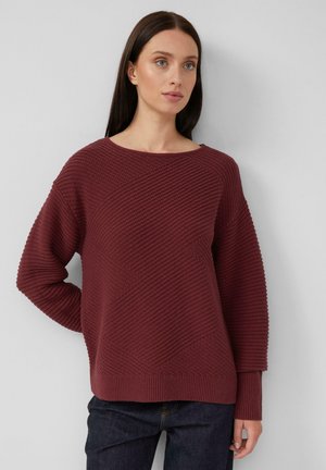 Vrouw met lang donker haar, gekleed in een gestructureerde bordeaux rode gebreide trui en donkere spijkerbroek, staande met één hand achter haar rug.