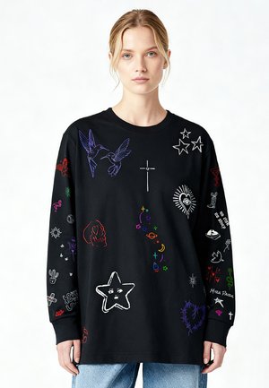 Jonge vrouw draagt een zwart oversized sweatshirt met kleurrijke geborduurde symbolen, waaronder sterren, vogels, harten, schedel en tekst, gecombineerd met een blauwe spijkerbroek.