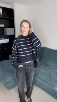 Maglione corto a righe blu navy e bianche con maniche lunghe, abbinato a jeans neri a vita alta e sneakers nere, contro un divano blu teal.