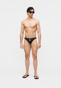 Man wearing black swim briefs with white logo, black flip-flops, and black sunglasses, standing against a plain light background.