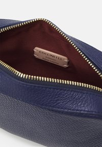 Borsa in pelle blu navy con zip dorata, con interni in tessuto bordeaux. L'etichetta recita "Coccinelle, Pelle Italiana Autentica."