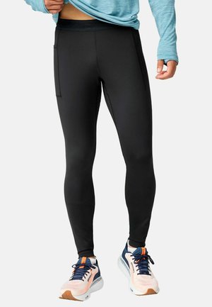 Person trägt schwarze sportliche Leggings mit Seitentaschen, ein hellblaues Langarmshirt sowie blaue und pfirsichfarbene Laufschuhe und steht auf weißem Hintergrund.