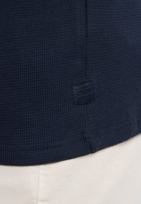 Námořnická modrá texturovaná trička s malou značkovou etiketu na lemu, v kombinaci se světlými kalhotami.